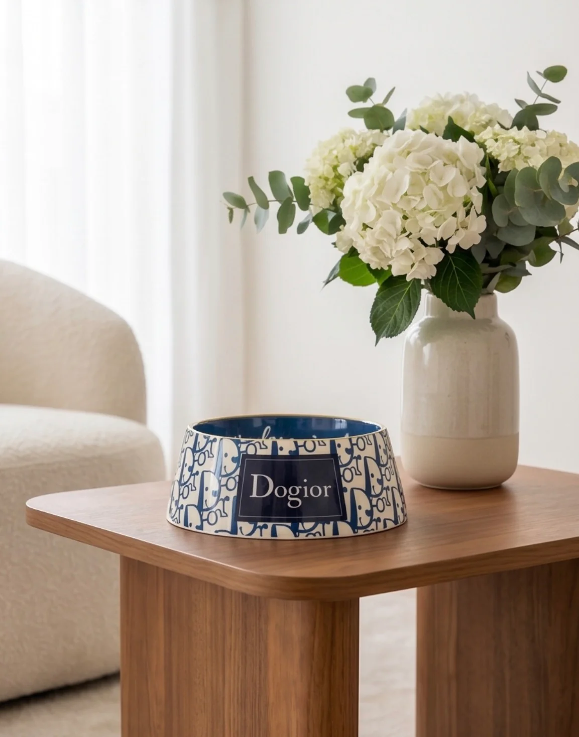 Blue-patterned pet bowl labeled 'Dogior' sits on a wooden table next to a vase of white hydrangeas and greenery in a bright living room.