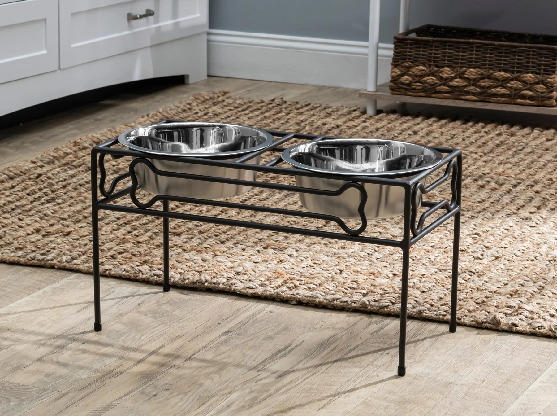 Two stainless steel pet bowls sit in a black metal stand on a woven rug in a kitchen setting.