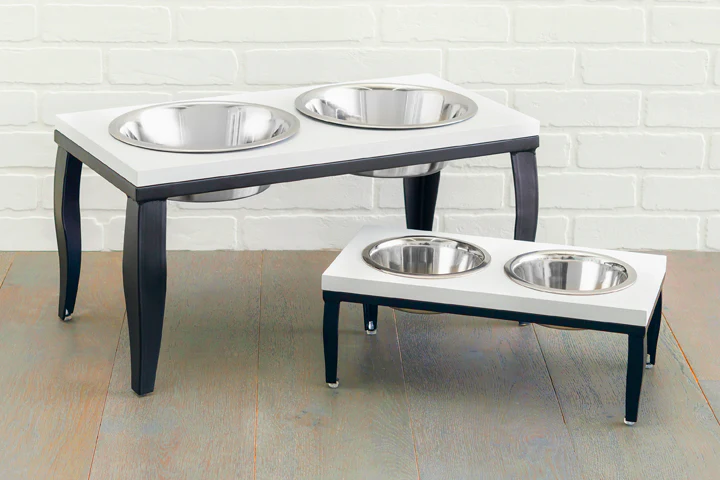 Two raised pet feeding stations with stainless steel bowls, a larger table and a smaller one, on a light wooden floor against a white brick wall.