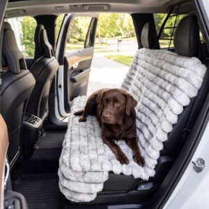 Brown dog lying on a thick gray fur blanket spread over the back seat of an open SUV, with front seats visible and a sunny suburban street outside the car.